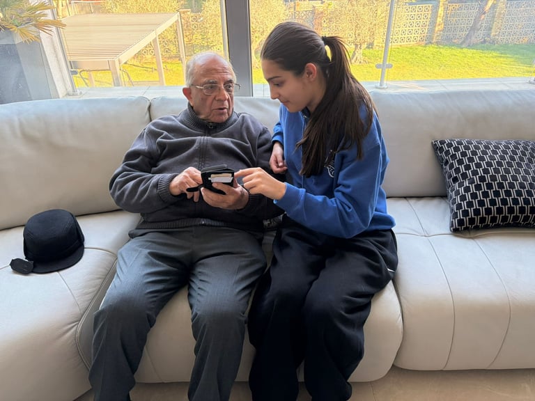 Elderly man and younger woman sit together on a beige sofa, holding and looking at a video game controller